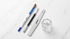Top-down view of professional DeEnti nail technician tools on a white marble surface, including a Kolinsky 16 acrylic nail brush, a dual-ended metal cuticle pusher, a 100-grit nail file, and a clear glass dappen dish.