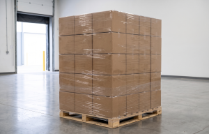 A wooden shipping pallet tightly stacked with 60 blank brown corrugated cardboard boxes wrapped in clear stretch film. The pallet sits on a polished concrete floor inside a clean, modern warehouse with a loading dock door visible in the background.