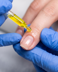 Professional nail technician wearing blue nitrile gloves applying hydrating cuticle oil to a client's natural nail to promote flexibility.